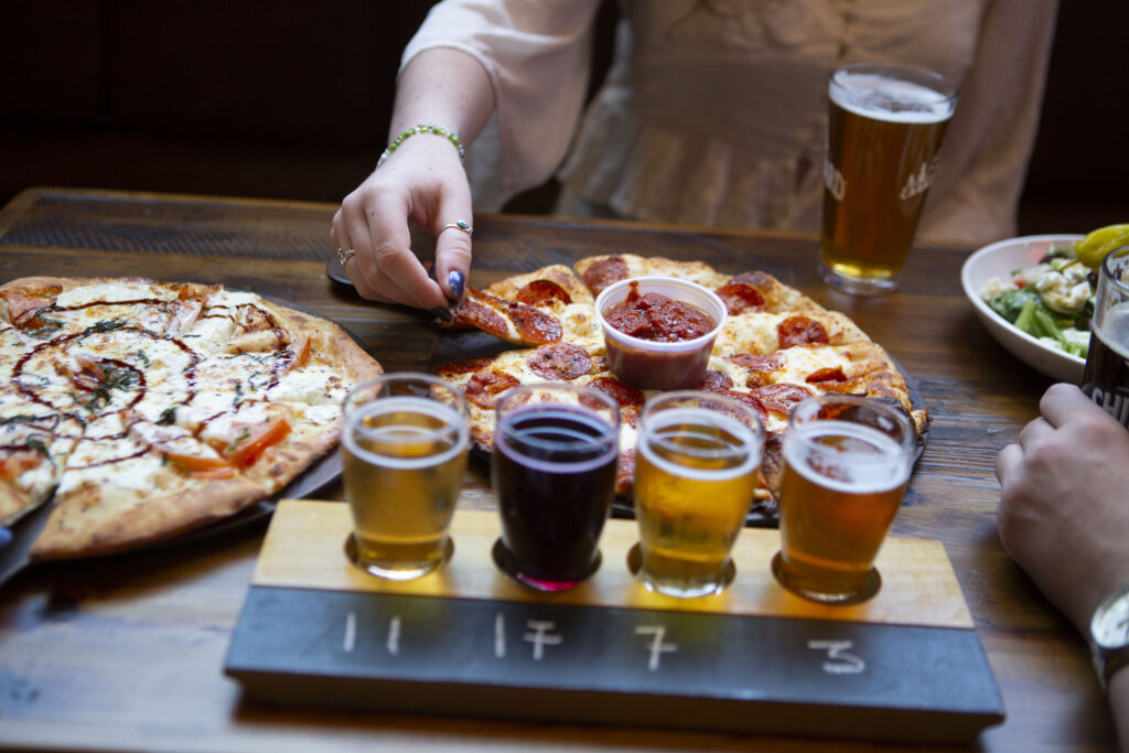 People sharing pizzas and a beer flight at a tabletop meal, one hand reaching for a slice over two large pepperoni pizzas and side dishes in a casual setting.
