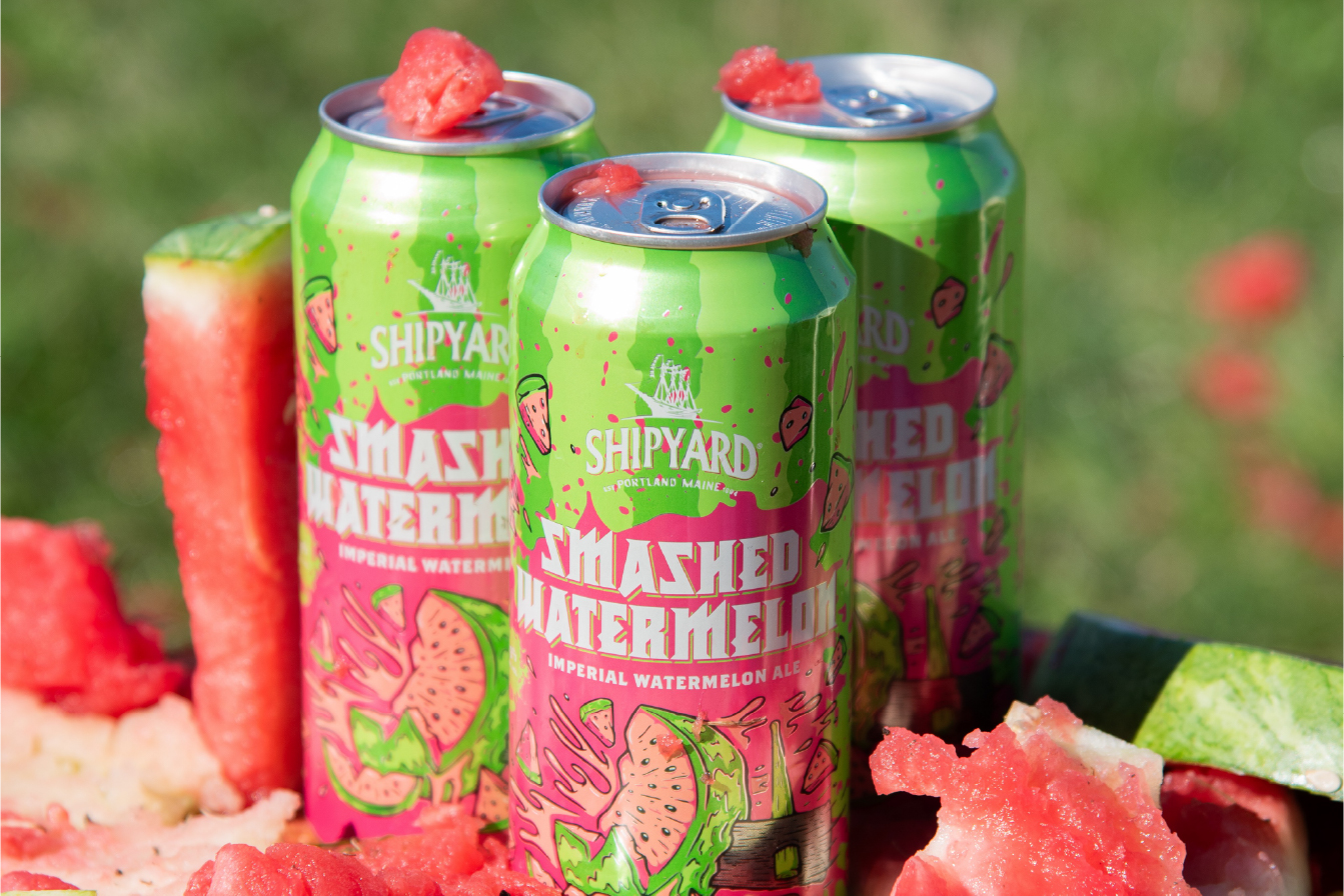 Three green cans of Shipyard Smashed Watermelon Imperial Watermelon Ale are surrounded by slices and chunks of fresh watermelon, set outdoors on a sunny day.