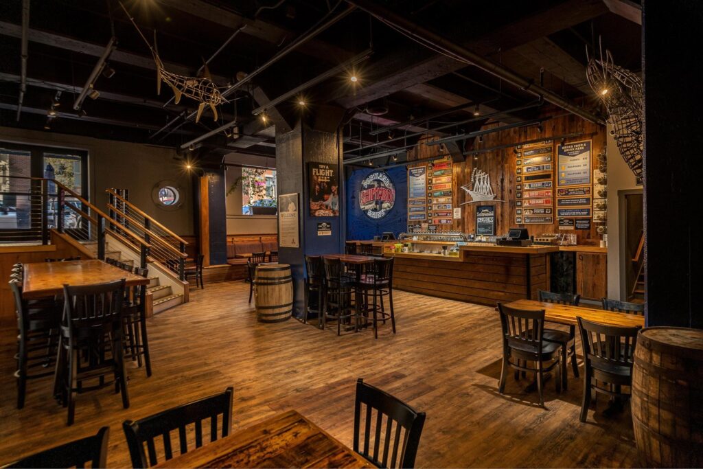 A cozy, rustic bar with wooden floors and furniture, a counter displaying colorful menus, posters on the walls, and warm ambient lighting. Stairs and large windows are visible in the background.
