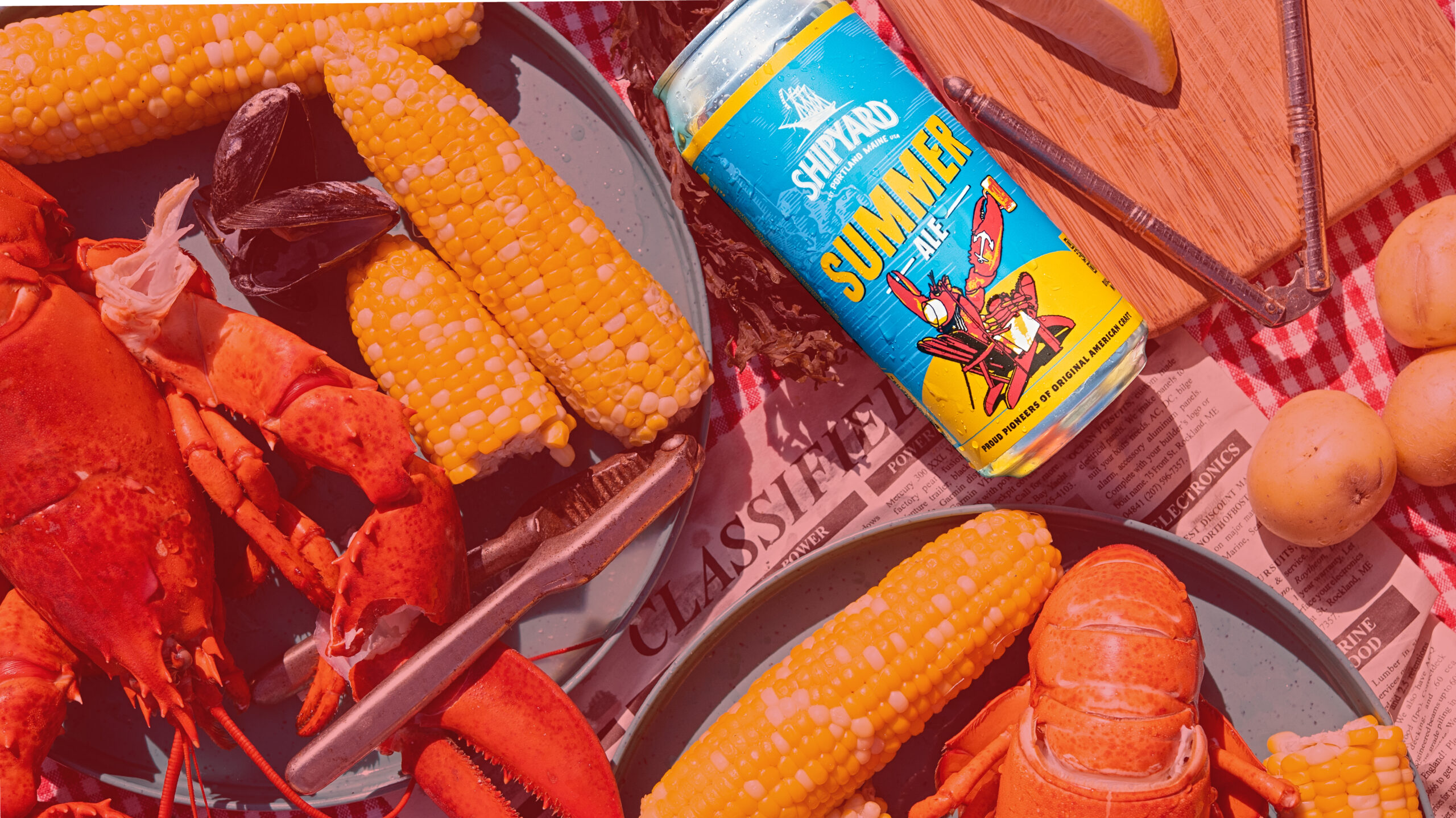 A picnic table with lobster, corn on the cob, mussels, potatoes, and a can of Shipyard Summer Ale on a blue plate over a red checkered cloth and newspaper.
