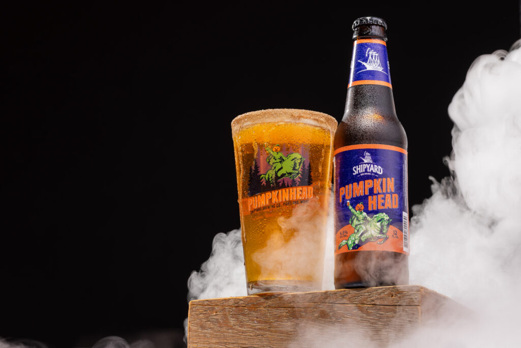 Bottle and glass of Shipyard Pumpkin Head beer on a wooden surface with a dark background and smoky fog
