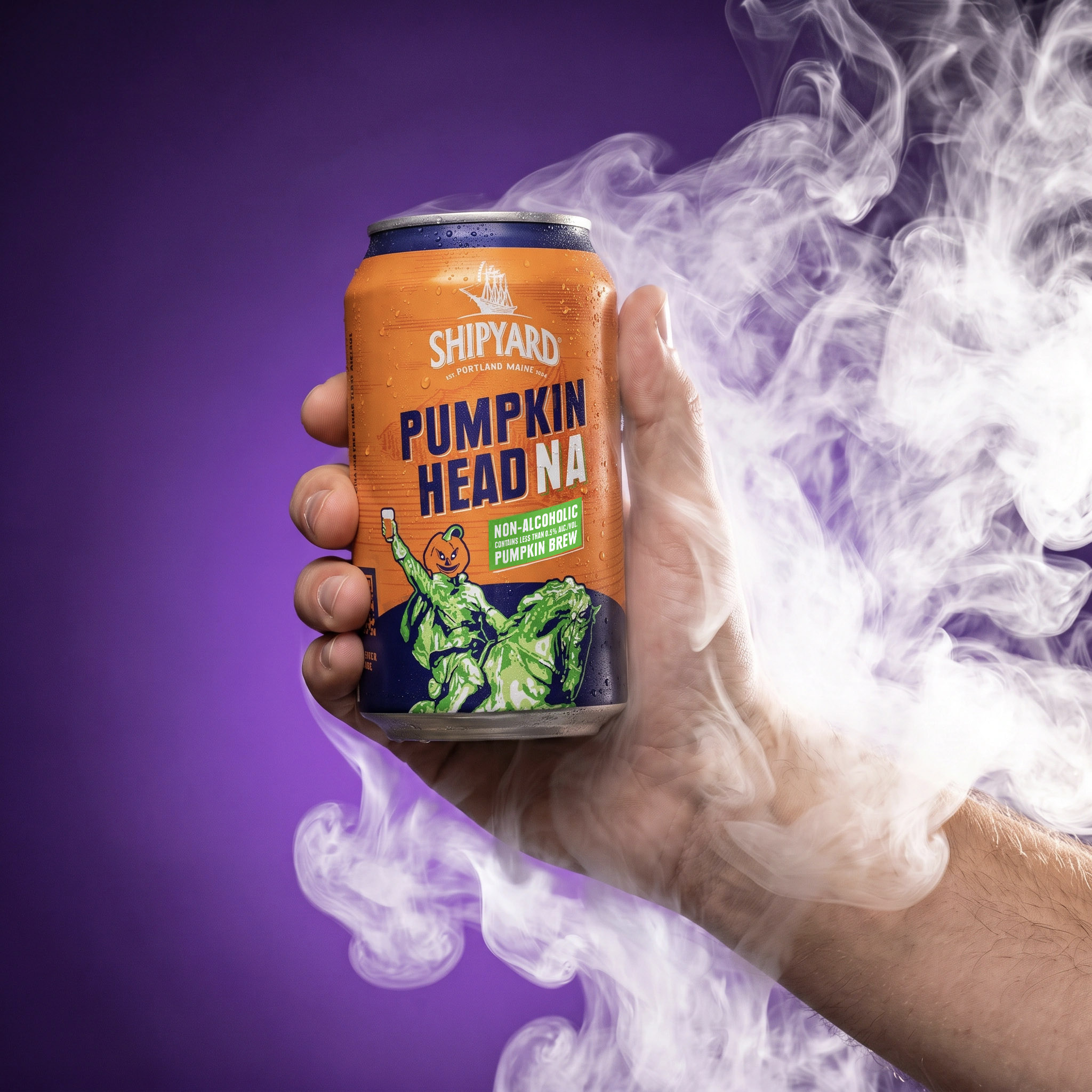 A hand holds a can of Shipyard Pumpkin Head NA non-alcoholic pumpkin brew against a purple background with white smoke swirling around.