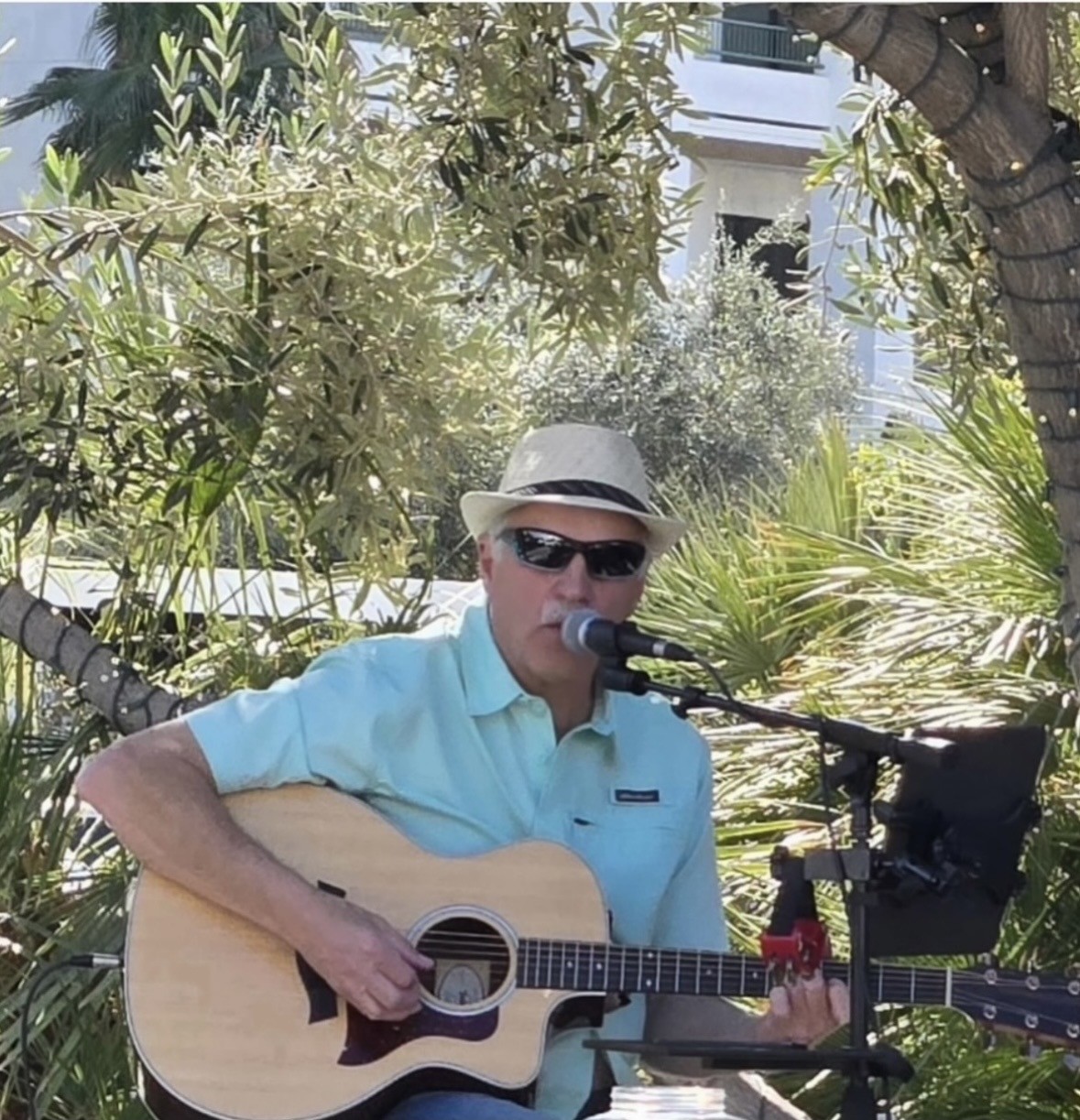 Man wearing a light blue shirt, sunglasses, and straw hat plays acoustic guitar and sings into a microphone outdoors among green trees and plants.